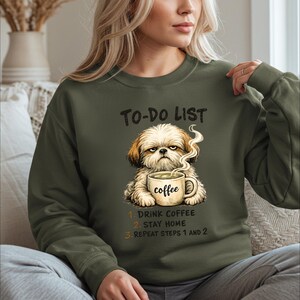 May include: An olive green crewneck sweatshirt featuring a graphic of a grumpy Shih Tzu dog holding a steaming mug labeled coffee. Above the dog is the text To-Do List, and below are the numbered steps: 1. Drink coffee, 2. Stay home, 3. Repeat steps 1 and 2.