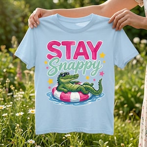 May include: A light blue t-shirt featuring a graphic of a green alligator wearing sunglasses and relaxing in a pink and white striped pool float. The text STAY Snappy is printed above the alligator in pink and light green, surrounded by yellow stars.