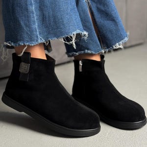 Könnte beinhalten: Ein Paar schwarze Wildleder-Stiefeletten mit abgerundeter Zehenpartie, flacher schwarzer Sohle und einer silberfarbenen, quadratischen Zierschnalle an der Außenseite. Die Stiefel verfügen über einen seitlichen Reißverschluss und eine glatte, matte Oberfläche.