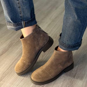 Könnte beinhalten: Ein Paar taupefarbene Wildleder-Stiefeletten mit abgerundeter Zehenpartie, einer dunkelbraunen Gummisohle mit strukturiertem Rand und einem vertikalen Reißverschluss an der Ferse. Die Stiefel werden zu einer blauen Jeans mit Umschlag getragen.