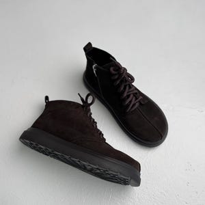 Könnte beinhalten: Ein Paar dunkelbraune Wildleder-Stiefeletten mit dicken Schnürsenkeln in Seiloptik, seitlichem Reißverschluss, einer Zugschlaufe an der Ferse und einer klobigen schwarzen Gummisohle mit strukturiertem Profil für Halt und Stabilität.