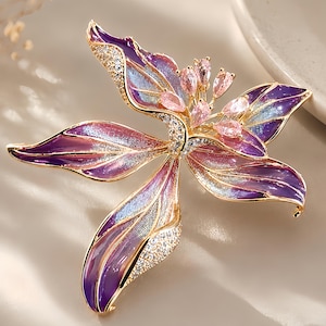Pode incluir: Um broche de flor em tom dourado com pétalas de esmalte roxo e azul com acabamento texturizado. O centro é adornado com vários cristais rosa claro em forma de pera e pequenos cristais transparentes ao longo das bordas das pétalas.
