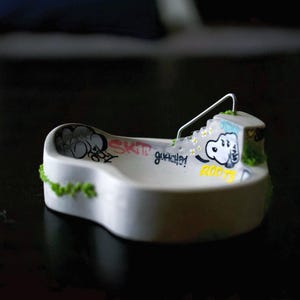 Puede incluir: Un bol de skate para fingerboard de hormigón blanco con una barandilla de metal, escaleras y arte de grafiti con el texto SKIT, guache y ROOTS. La estructura está decorada con pequeños parches de musgo artificial verde en los bordes exteriores.