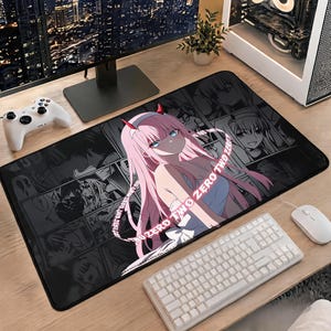 May include: A rectangular desk mat featuring a black and white manga-style collage background with a central colored illustration of an anime character with long pink hair and red horns, accompanied by the repeating text ZERO TWO ZERO TWO.