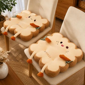 May include: Two square plush seat cushions shaped like toast with rounded edges. The cushions are light beige with brown textured sides, featuring embroidered faces, small orange carrot-like arms, and the text "I AM TOAST" on the surface.