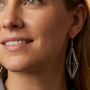 May include: A pair of silver-toned geometric earrings featuring a multi-faceted, elongated diamond shape constructed from thin metal rods. The earrings hang from simple hooks and have a polished, metallic finish that reflects light.