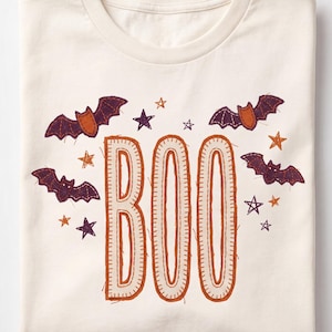 Puede incluir: Una camiseta de color crema con la palabra BOO en letras grandes de color naranja con un contorno estilo costura. El diseño incluye murciélagos voladores de color morado oscuro y naranja y varias estrellas dispersas alrededor del texto.