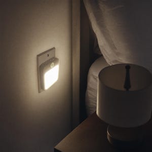 May include: A square, white motion-sensor night light plugged into a wall outlet. The device features a flat, illuminated front panel emitting a warm, bright glow, with a small circular sensor positioned at the top center of the light housing.