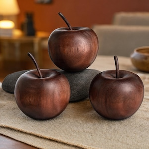 May include: Three smooth, dark brown wooden apple sculptures with visible wood grain patterns and thin, curved stems. The apples are rounded and polished, featuring a deep mahogany tone that highlights the natural texture of the wood material.