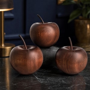 May include: Three decorative apple sculptures carved from dark brown wood, each featuring a smooth, polished surface with visible natural grain patterns and a slender, upright wooden stem attached to the top of each rounded fruit-shaped object.