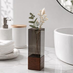 May include: A square-shaped vase featuring a dark grey translucent acrylic top section and a solid dark brown wooden base. The vase contains a single stem of green eucalyptus leaves and a small sprig of dried white flowers.