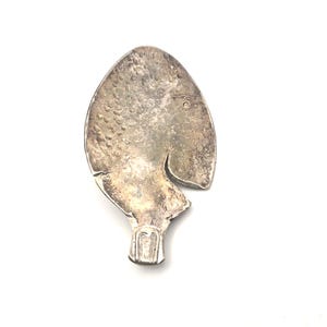 May include: A silver-toned metal pendant shaped like a fish with a textured scale pattern on the body, a small circular eye, and a defined tail section. The surface has a weathered, tarnished appearance with visible dark oxidation and scratches.
