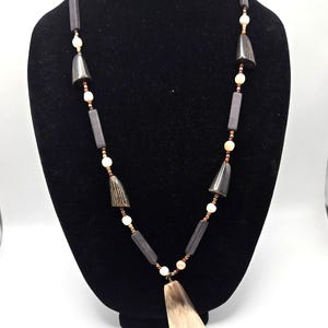 May include: A necklace featuring a large, tapered, light-colored horn pendant suspended from a strand of dark brown rectangular wooden beads, small cream-colored round beads, and dark, cone-shaped horn accents arranged in a repeating pattern.