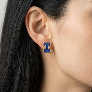 May include: A rectangular stud earring featuring a deep blue stone set in a gold-toned frame. The centre of the blue stone displays two small, symmetrical gold-toned teardrop or wing-shaped embellishments positioned side by side in the middle.