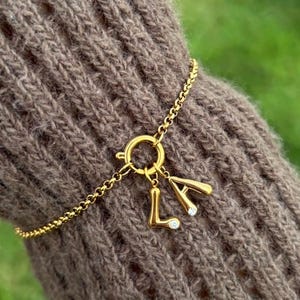 Könnte beinhalten: Ein goldfarbenes Kettenarmband mit einem runden Federringverschluss. Zwei goldfarbene Buchstabenanhänger, L und A, sind am Verschluss befestigt. Jeder Buchstabenanhänger hat einen kleinen, klaren, runden Edelstein an der Unterseite.