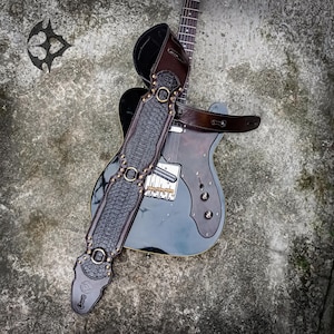 Pode incluir: Uma correia de guitarra em couro castanho escuro com um painel central preto texturizado, anéis de metal cor de latão e rebites. A correia está posicionada sobre uma guitarra elétrica preta com um escudo de madeira escura e ponte cromada.