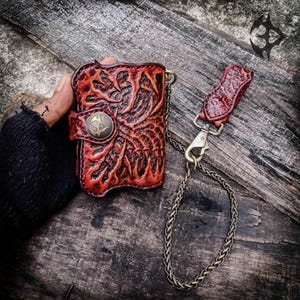 May include: A red leather wallet featuring a deeply embossed, organic, root-like pattern and a central brass-toned snap button closure. The wallet is attached to a matching leather belt loop tab by a long, thin, silver-toned metal chain.