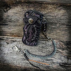 May include: A dark purple and black textured leather pouch featuring a lace-up side detail, silver-toned metal rivets, and a central circular metal emblem. A silver-toned metal chain with a lobster clasp and a thin leather cord is attached to the side.