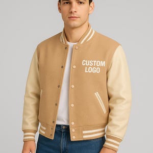 May include: A tan varsity jacket featuring cream-colored sleeves, a ribbed collar, cuffs, and hem with white stripes. The jacket has a snap-button front closure, two side pockets, and the text CUSTOM LOGO printed in white on the left chest area.
