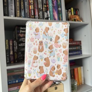 May include: A clear protective tablet case featuring a repeating pattern of brown capybaras engaged in various activities, including gardening, drinking tea, and bathing, surrounded by colourful flowers, strawberries, and small household items.