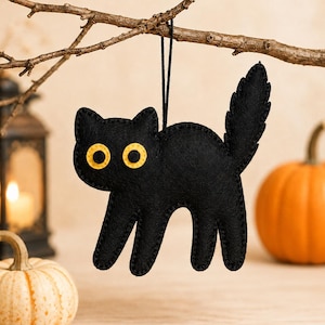 Puede incluir: Un adorno de fieltro negro con forma de gato, con cola puntiaguda y lomo arqueado, que presenta dos ojos redondos amarillos con centros negros. El gato está suspendido por un cordel negro fino y tiene costuras visibles en los bordes.