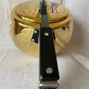 May include: A gold-colored pressure cooker with a black handle and lid. The cooker has a shiny, reflective surface and a black pressure release valve. The handle is attached with silver rivets. The text "A Symbol of Quality" is visible on the lid.
