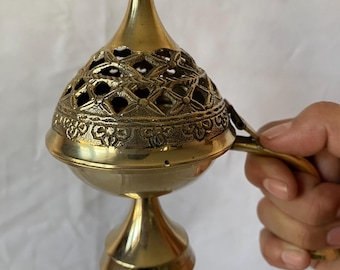 May include: A brass incense burner with a decorative, pierced lid and a handle. The burner has a round base and a conical top. The intricate design on the lid features floral and geometric patterns. The overall color is a warm, antique gold.