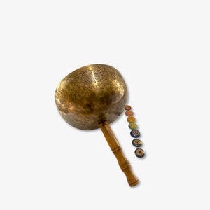 Puede incluir: Un cuenco de canto grande, redondo y de latón con un mango de madera. El cuenco está grabado con caracteres sánscritos y está rodeado de siete piedras de chakra.