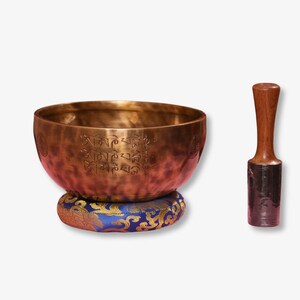May include: A traditional Tibetan singing bowl with a wooden striker. The bowl is made of brass and has a gold and brown patina. It is decorated with Tibetan script and sits on a blue and gold cushion.