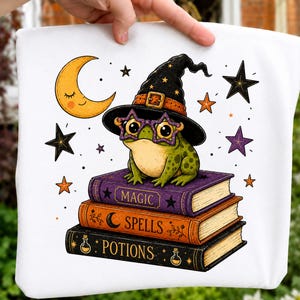 Op de afbeelding: Een wit stoffen item met een afbeelding van een groene kikker met een zwarte heksenhoed en stervormige bril, zittend op een stapel van drie boeken met de tekst Magic, Spells en Potions, omringd door een gele maansikkel en zwarte sterren.