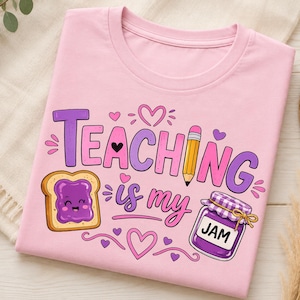 以下が含まれることがあります： 「TEACHING is my JAM」という文字がプリントされたライトピンクのTシャツ。紫色のジャムが塗られた笑顔のトースト、黄色い鉛筆、紫色のジャム瓶、そしてピンクと紫の小さなハートが描かれたグラフィックデザインが特徴です。