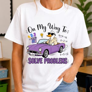 May include: A white t-shirt featuring a graphic of a duck wearing sunglasses and a hoodie, driving a purple convertible car. The text reads "On My Way To SOLVE PROBLEMS" with math equations, a calculator, and a lightbulb icon surrounding the duck.