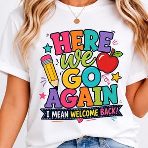 May include: A white short-sleeve t-shirt featuring a colorful graphic with the text "HERE WE GO AGAIN I MEAN WELCOME BACK!" alongside illustrations of a yellow pencil, a red apple, and small stars in shades of pink, teal, and yellow.