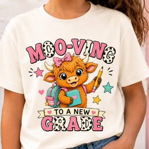May include: A cream-colored t-shirt featuring a graphic of a fluffy brown highland cow wearing a pink polka-dot bow. The cow holds a pencil and a book, with a backpack nearby. The text reads MOO-VING TO A NEW GRADE in pink and cow-print letters.