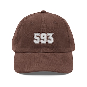 May include: A brown corduroy baseball cap featuring the number 593 embroidered in white with a dark outline on the front centre. The hat has a structured crown, a curved brim, and a button at the top, showcasing a textured fabric finish.