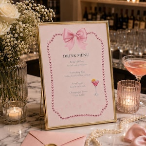 Peut inclure: Un menu de boissons rectangulaire dans un cadre doré, orné d'un nœud rose et d'une bordure de perles. Le texte indique DRINK MENU, Bride's Bellini, Something Blue, Rosé Spritz et Champagne Tower, avec une illustration de verre à cocktail.