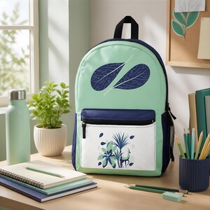 May include: A mint green and navy blue backpack featuring a dark blue leaf pattern on the upper section and a white front pocket displaying a graphic illustration of tropical plants and a small deer in shades of blue and green.