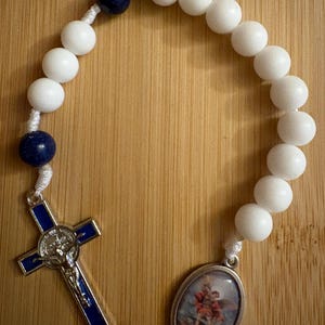 May include: A rosary featuring white spherical beads, two dark blue spherical beads, and a silver-toned crucifix with blue enamel inlay. An oval-shaped metal pendant depicting a religious figure in red and brown robes is attached to the white cord.