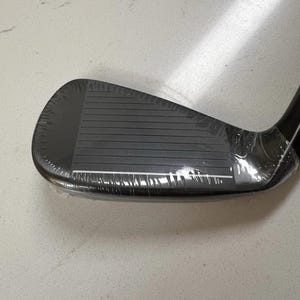 Puede incluir: Una cabeza de palo de golf de hierro en color negro mate con una serie de l&iacute;neas horizontales en la cara. La cabeza del palo est&aacute; parcialmente envuelta en una pel&iacute;cula pl&aacute;stica protectora transparente, mostrando un acabado liso y curvo.