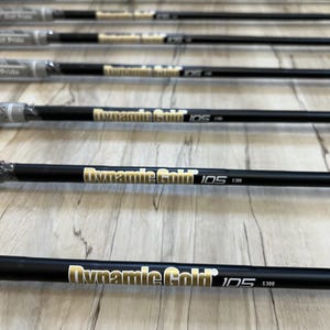 Puede incluir: Un conjunto de siete varas de acero negro para palos de golf dispuestas en paralelo. Cada vara presenta un texto en dorado y blanco que dice Dynamic Gold 105 S300. Los extremos superiores tienen empu&ntilde;aduras de goma gris, algunas envueltas en pl&aacute;stico.