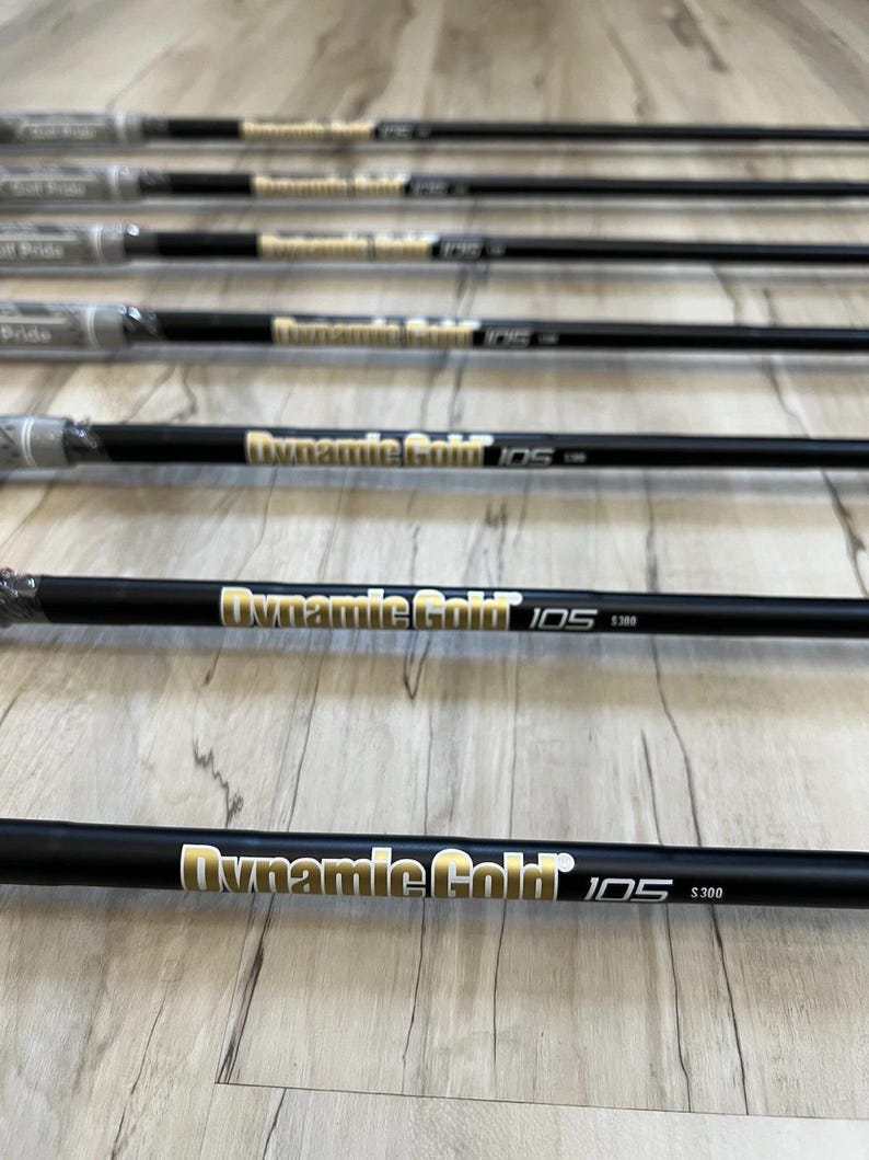 Puede incluir: Un conjunto de siete varas de acero negro para palos de golf dispuestas en paralelo. Cada vara presenta un texto en dorado y blanco que dice Dynamic Gold 105 S300. Los extremos superiores tienen empu&ntilde;aduras de goma gris, algunas envueltas en pl&aacute;stico.