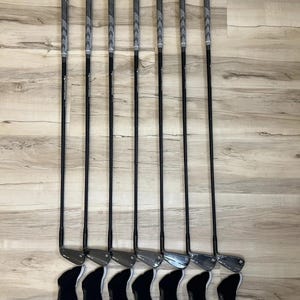 Puede incluir: Un juego de siete hierros de golf con varillas negras y empu&ntilde;aduras grises texturizadas, cada uno con una funda protectora negra. Los palos tienen cabezas met&aacute;licas con un acabado liso, dispuestos en fila vertical con sus fundas debajo.