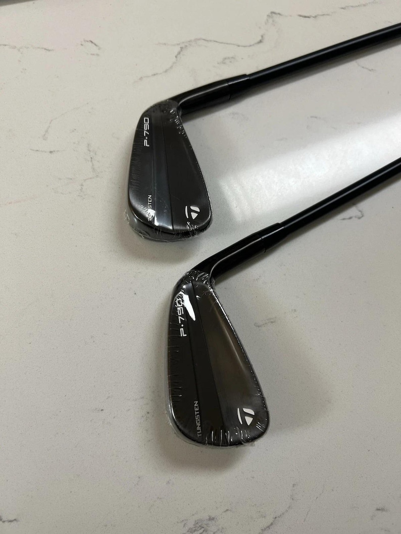 Puede incluir: Dos hierros de golf TaylorMade P-790 negros con varillas negras. Cada cabeza de palo presenta el texto P-790 y TUNGSTEN impreso en blanco, junto con el logotipo de TaylorMade. Las cabezas de los palos est&aacute;n envueltas en film pl&aacute;stico transparente.