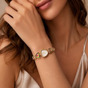 May include: A gold-tone watch featuring a triangular white face and a bracelet composed of alternating gold-tone links and iridescent, multi-colored triangular stones. The watch has a delicate, link-style band that secures around the wrist.