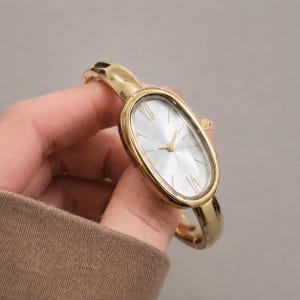 Op de afbeelding: Een goudkleurig armbandhorloge met een ovale kast en een gladde, gepolijste metalen band. De wijzerplaat is zilverkleurig met dunne goudkleurige uurmarkeringen, inclusief Romeinse cijfers op twaalf en zes uur, en goudkleurige wijzers.