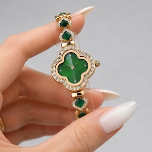 May include: A gold-tone wristwatch featuring a clover-shaped face with a deep green dial, surrounded by a border of clear crystals. The watch band consists of gold-tone links, each set with a square green stone and accented with clear crystals.