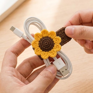 May include: A white USB charging cable secured with a brown and yellow crocheted sunflower cord organizer. The sunflower features a dark brown center with bright yellow petals, attached to a brown crocheted strap that wraps around the coiled cable.