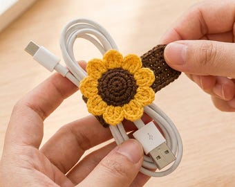 May include: A white USB charging cable secured with a brown and yellow crocheted sunflower cord organizer. The sunflower features a dark brown center with bright yellow petals, attached to a brown crocheted strap that wraps around the coiled cable.