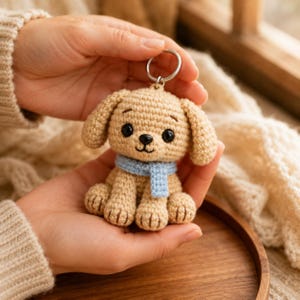 Può includere: Un portachiavi a forma di cane realizzato all'uncinetto in color beige con una sciarpa azzurra, occhi in plastica nera e naso ricamato. Il piccolo peluche è fissato a un anello in metallo argentato, con una trama in filato dettagliata.