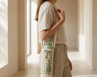 May include: A cream-colored crocheted water bottle holder with a long shoulder strap. The holder features a grid-like pattern, a decorative green tassel, and two small wooden beads tied with a green string near the top opening.